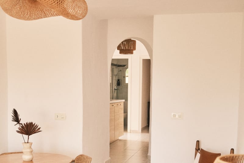 Sunlit dining nook in the apartment with a round wooden table, woven pendant light, and an arched doorway leading deeper into the unit.