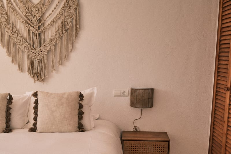 Second bedroom with white linen, dark wooden side table with rattan front, and a large macrame hanging above the bed.