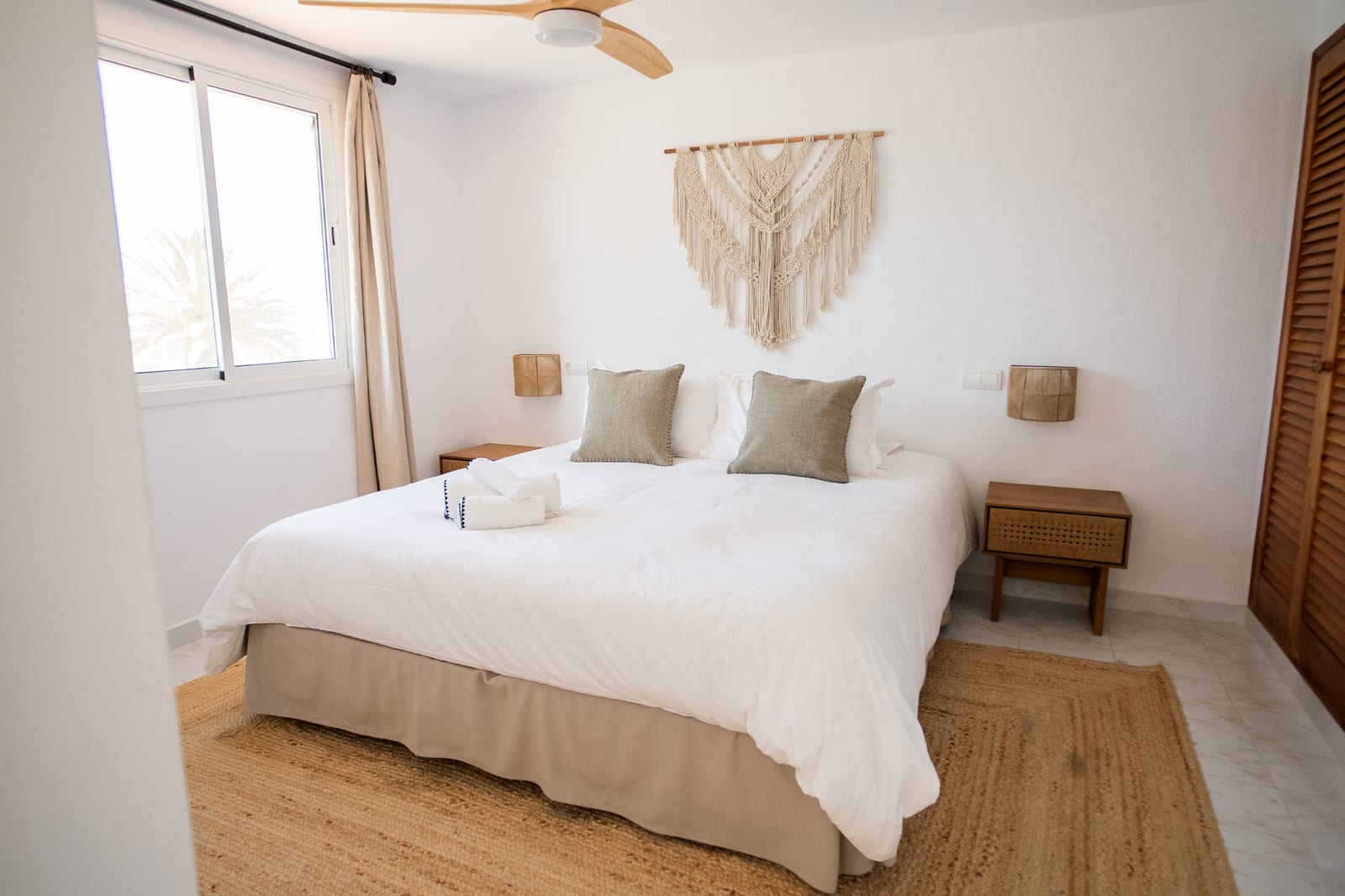 Whitewashed apartment bedroom with a king bed, a hanging macrame wall piece, ceiling fan, linen curtains, and a large jute rug under bare feet.