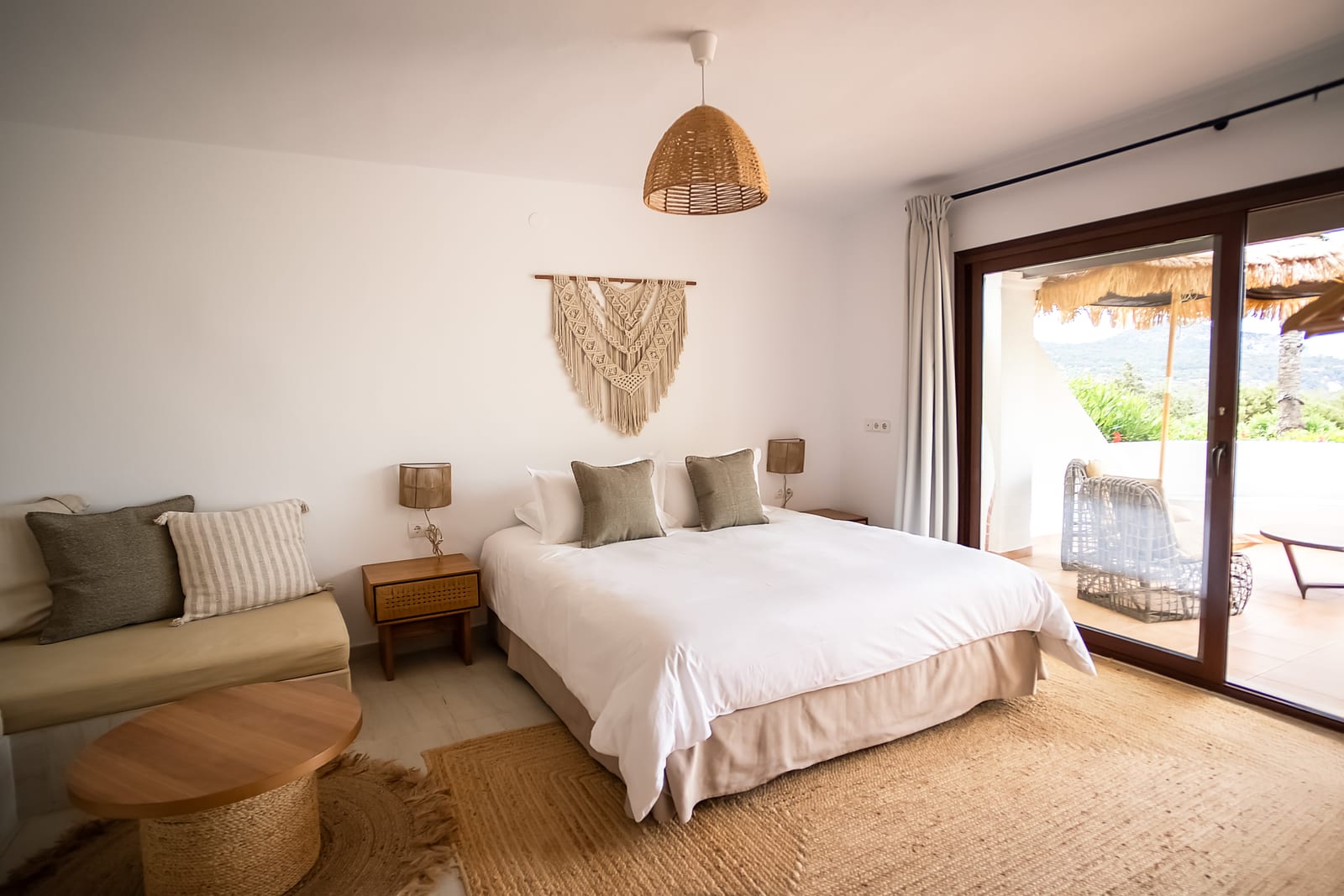 Light, simple bedroom with a king bed, jute rug, small cream sofa, and sliding doors opening onto a shaded pergola terrace with rattan chairs.
