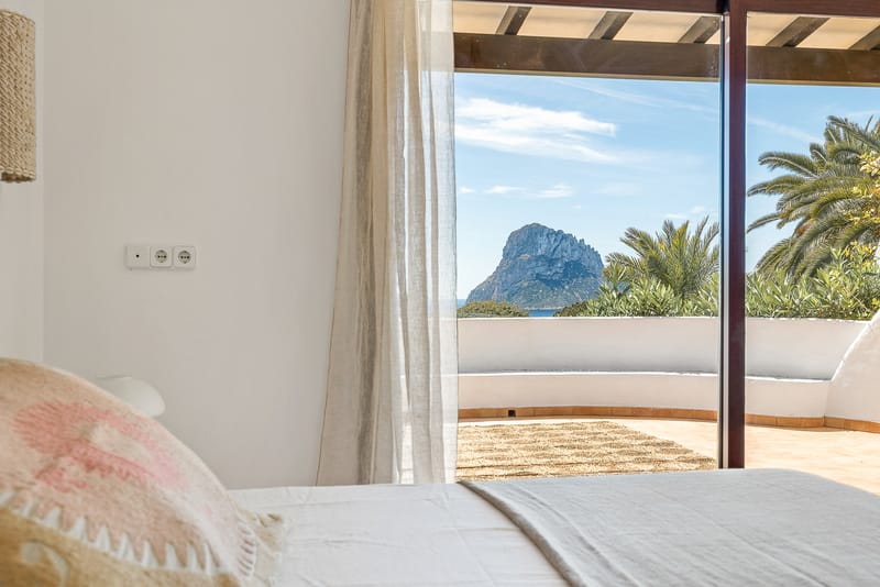 Bedroom doorway view of Es Vedrà rising from the sea, framed by palm fronds and white curtains.