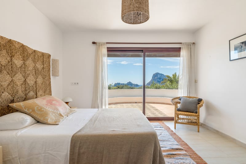 Closer angle of the suite bed showing a printed cushion, terracotta runner, and a rattan armchair by the balcony door.