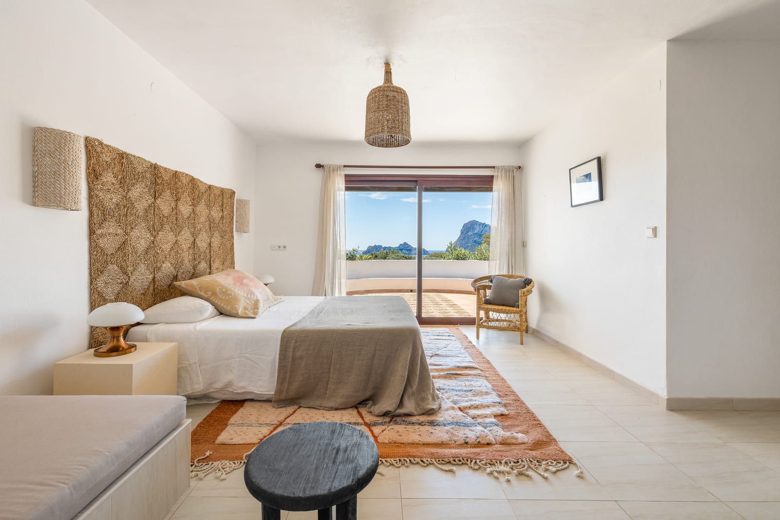 Bright open bedroom with a king bed, woven headboard, terracotta rug, and floor-to-ceiling sliding doors revealing the Es Vedrà sea rock and the Mediterranean beyond.