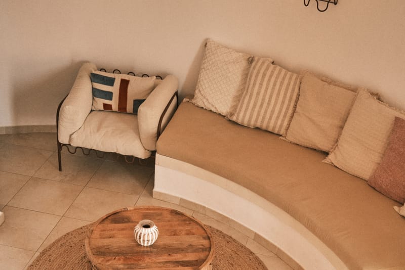 Curved built-in sofa lined with linen cushions, encircling a low round jute rug and wooden coffee table.