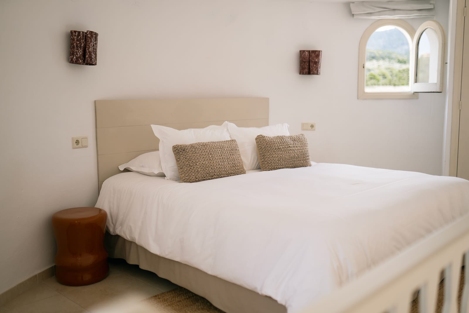 Calm white bedroom with a low headboard, crisp white linen, jute pillows, and a small arched window cut into a thick stucco wall looking out toward green hills.